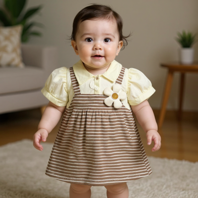 Baby wearing a yellow blouse and brown striped pinafore dress with a flower detail indoors.