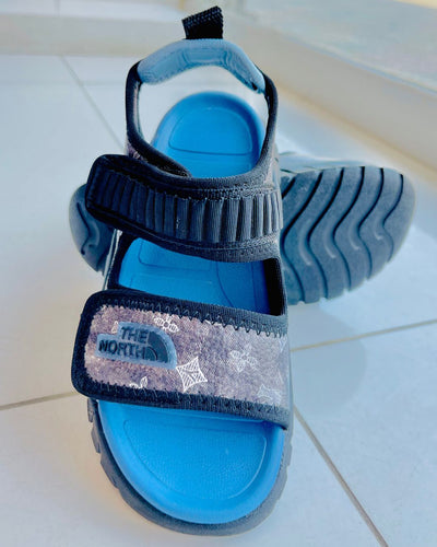 Blue sandal with black strap featuring The North Face logo on a tiled floor.