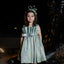 Young girl in a green dress with a decorative headband standing against a dark background.