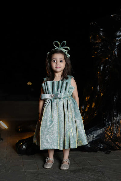 Young girl in a green dress with a decorative headband standing against a dark background.