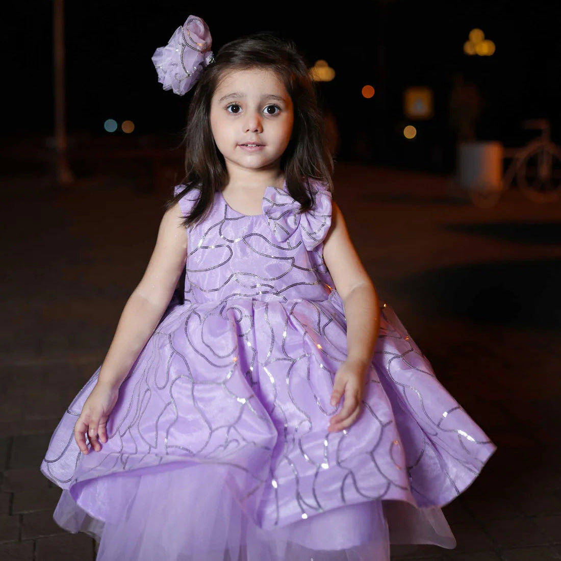 Young girl in a purple dress with a flower in her hair, standing outdoors at night.