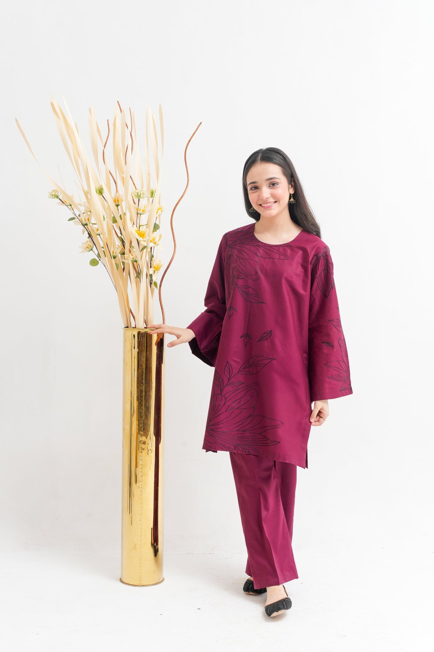Woman in a maroon outfit standing next to a tall gold vase with decorative branches on a white background
