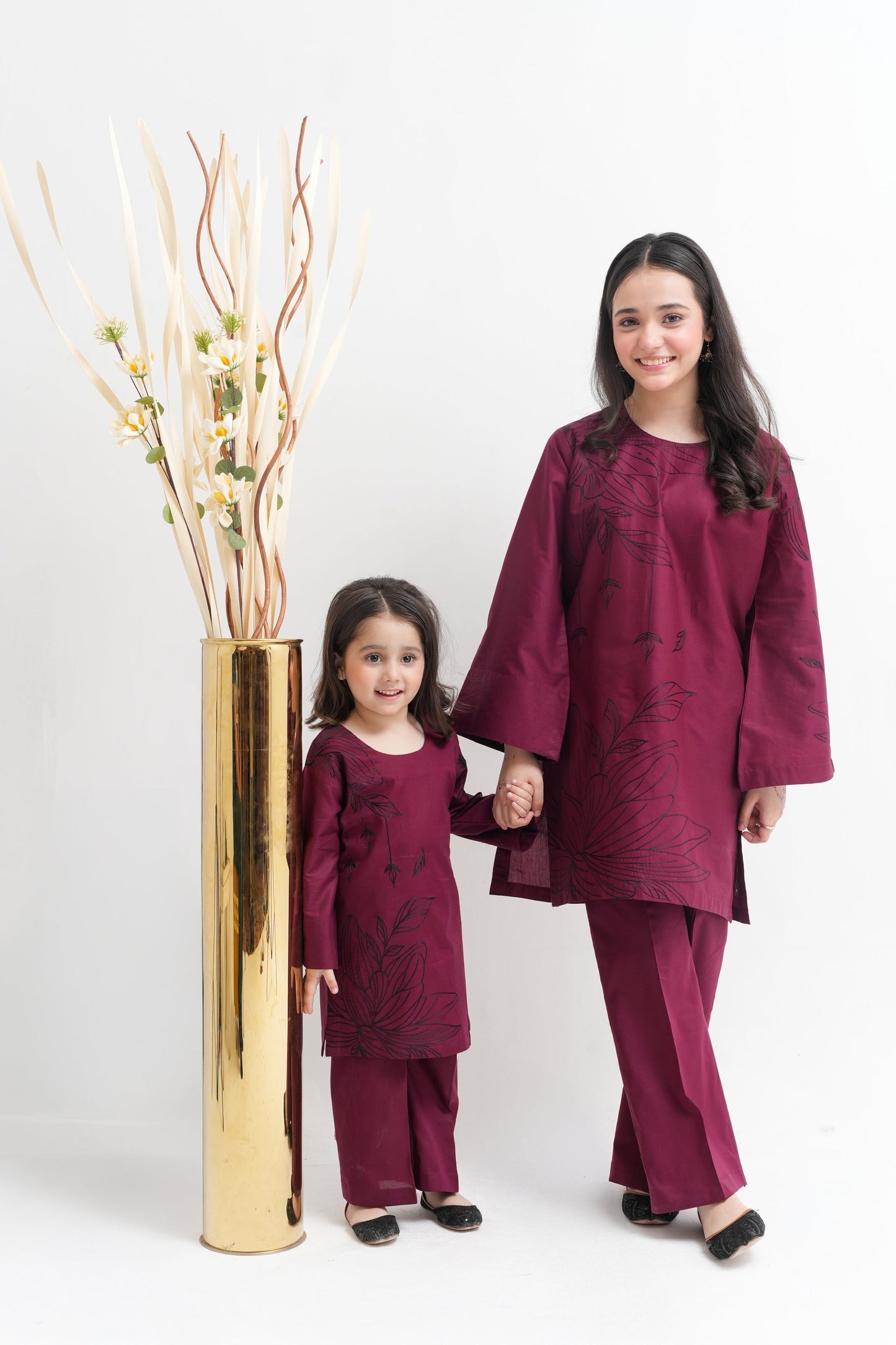 Woman and child in matching maroon outfits standing next to a gold vase with decorative branches on a white background