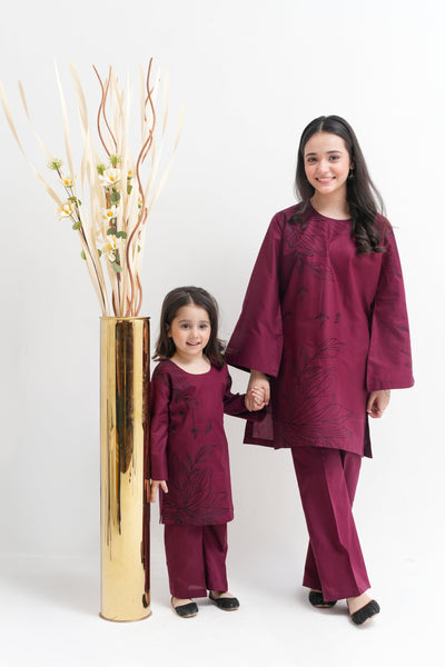 Woman and child in matching maroon outfits standing next to a gold vase with decorative branches on a white background