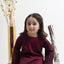 Young girl in a burgundy outfit sitting on a white surface with decorative elements.