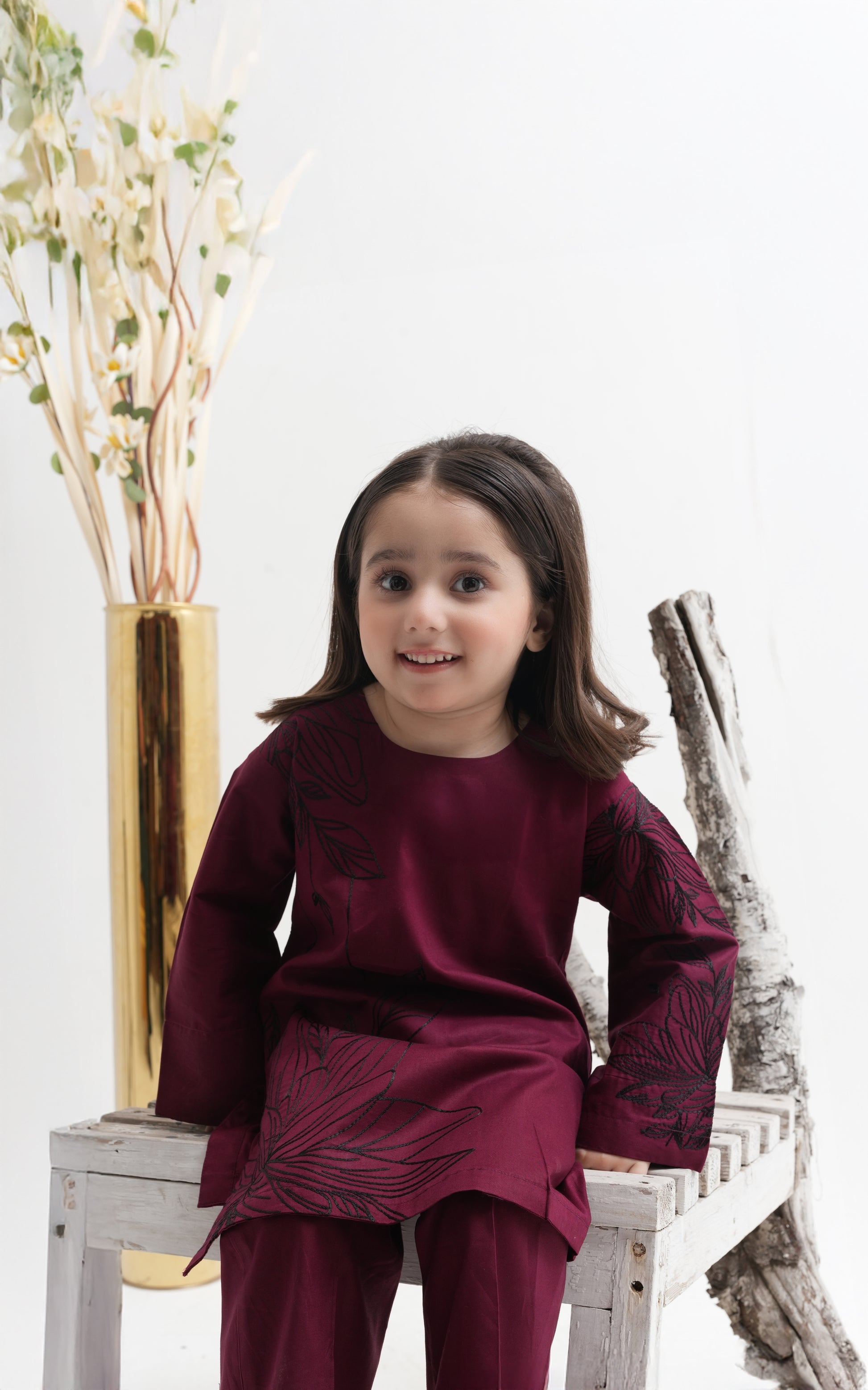 Young girl in a burgundy outfit sitting on a white surface with decorative elements.