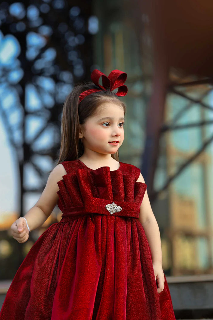 Young girl in a red dress with a large bow in her hair, standing outdoors.
