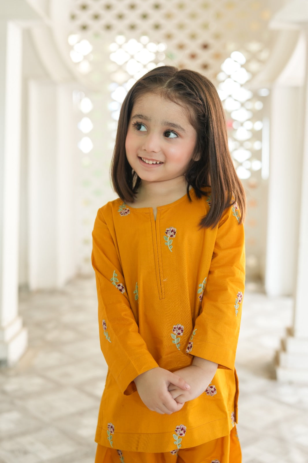 Young girl wearing a mustard yellow traditional outfit with floral patterns indoors.